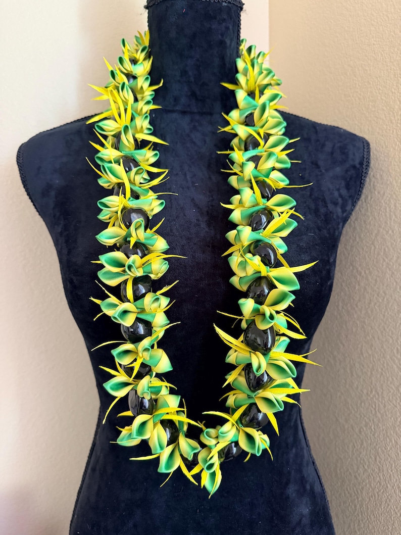 Pu&ograve; includere: Una vivace collana lei con petali a nastro verdi e gialli alternati e perline nere. La lei &egrave; esposta su un manichino nero, mettendo in risalto il suo design colorato e il tema tropicale.