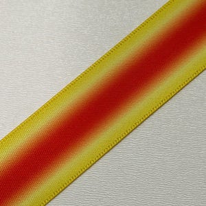 Pu&ograve; includere: Primo piano diagonale di un nastro con un design sfumato. Il nastro passa da bordi gialli a un centro rosso vibrante. I bordi hanno un aspetto strutturato e tessuto, e lo sfondo &egrave; una superficie bianca strutturata.