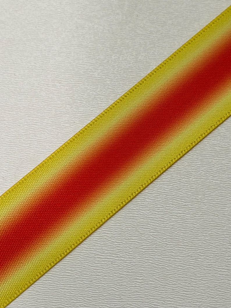 Pu&ograve; includere: Primo piano diagonale di un nastro con un design sfumato. Il nastro passa da bordi gialli a un centro rosso vibrante. I bordi hanno un aspetto strutturato e tessuto, e lo sfondo &egrave; una superficie bianca strutturata.
