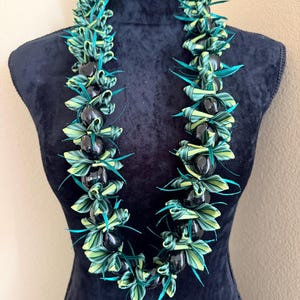 Pu&ograve; includere: Una vivace collana lei esposta su un manichino nero. La lei presenta perline nere alternate e foglie intrecciate verdi e gialle con accenti turchese. La collana &egrave; una ghirlanda hawaiana tradizionale, spesso indossata per le celebrazioni.