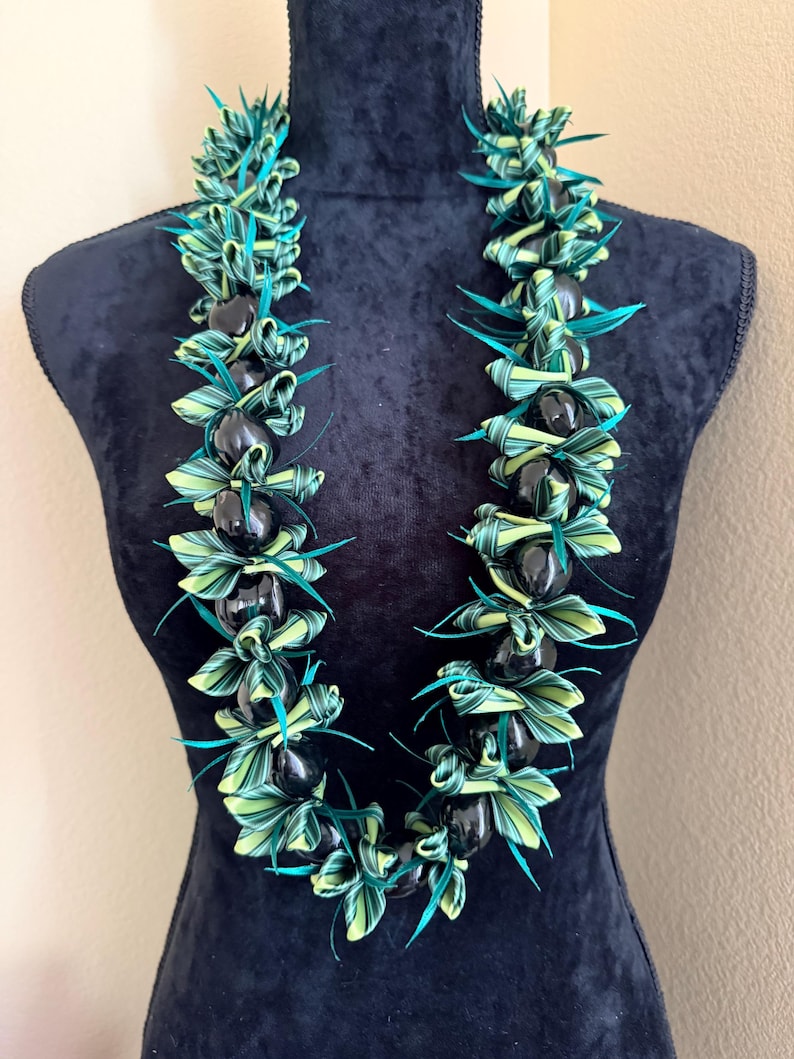Pu&ograve; includere: Una vivace collana lei esposta su un manichino nero. La lei presenta perline nere alternate e foglie intrecciate verdi e gialle con accenti turchese. La collana &egrave; una ghirlanda hawaiana tradizionale, spesso indossata per le celebrazioni.