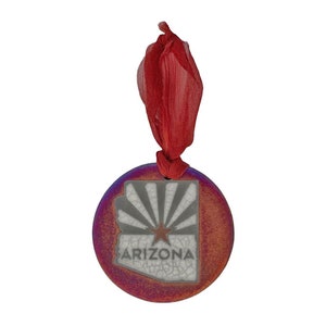 Può includere: Un ornamento rotondo in ceramica con una glassa rossa, viola e arancione. L'ornamento presenta un contorno bianco dello stato dell'Arizona con una stella rossa e la parola "Arizona" in lettere nere. L'ornamento ha un nastro rosso per appenderlo.