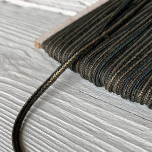 May include: A spool of dark green cord with a subtle metallic gold thread woven throughout. The cord is tightly wound around a rectangular cardboard base, with a single strand extending outward, showcasing the braided texture and color.