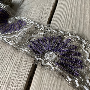 May include: A close-up of a beaded trim with a purple floral design. The trim is made of silver and white beads and is on a white background.
