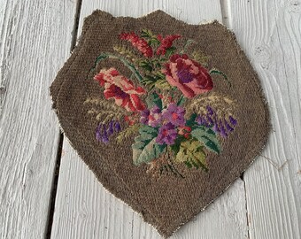 May include: A vintage needlepoint floral design featuring a bouquet of pink, red, and purple flowers with green leaves on a brown background.