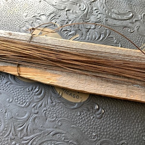 May include: A bundle of thin, brown wire is neatly arranged on a weathered wooden plank. The wood has a natural, aged appearance. The wire is a rich, warm brown color. The background is a textured, dark gray surface with a floral pattern.