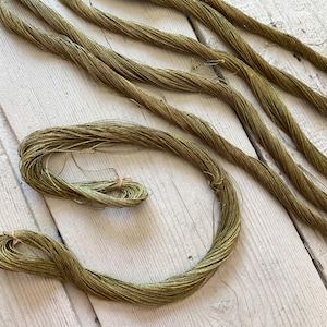 May include: Olive green embroidery floss, arranged on a light wood surface. Several strands are straight, while one is coiled and secured with a small pink rubber band. The floss has a slight sheen.
