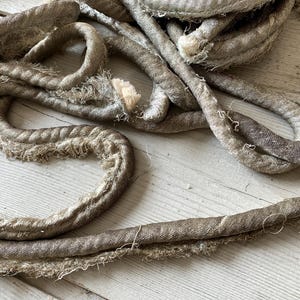 May include: Close-up of several strands of thick, textured rope in varying shades of gray and beige. The rope appears to be made of fabric, with frayed edges and a slightly worn appearance. The rope is arranged in a tangled, overlapping pattern.