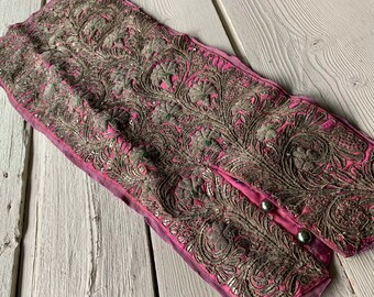 May include: A pink fabric trim with silver embroidery featuring a floral pattern. The trim has two silver buttons.