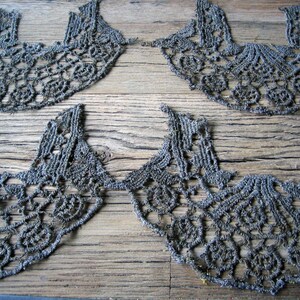 May include: Several gray lace collars are arranged on a weathered wooden surface. The collars have a scalloped edge and intricate floral and geometric patterns. The lace appears to be handmade, with visible texture and detail.