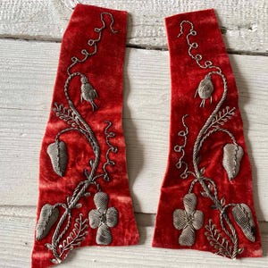May include: Two red velvet fabric pieces with silver embroidered floral designs. The embroidery includes leaves, flowers, and vines. The fabric has a soft, textured appearance, and the embroidery is detailed and intricate.