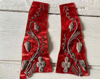 May include: Two red velvet fabric pieces with silver embroidered floral designs. The embroidery includes leaves, flowers, and vines. The fabric has a soft, textured appearance, and the embroidery is detailed and intricate.