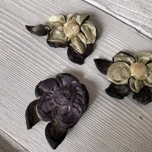 May include: Three handmade fabric flower embellishments. Two flowers are gold and black, and one is purple and black. The flowers have layered petals and are arranged on a light wood grain surface.
