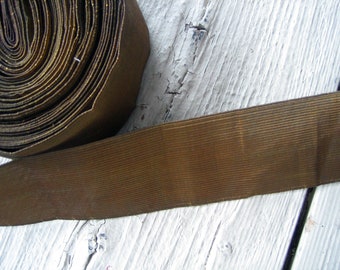May include: A roll of brown grosgrain ribbon with gold thread woven into the fabric. The ribbon is rolled up and the end is lying flat on a white wooden surface.