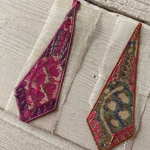 May include: Two decorative fabric pieces with a geometric design. One is a deep magenta with gold and purple accents, the other is orange with blue and green details. Both are on a beige mesh backing.