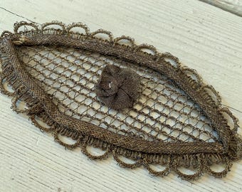 May include: An oval-shaped, brown decorative piece with a woven net design. The edges are trimmed with a scalloped border. A small, textured, brown fabric flower is centered on the net. The piece is likely vintage or antique.