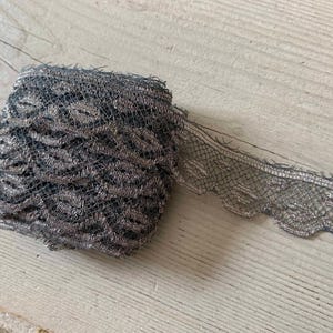 May include: A roll of gray lace with a scalloped edge. The lace has a metallic sheen and a delicate, open weave pattern. The roll is sitting on a light-colored wooden surface.