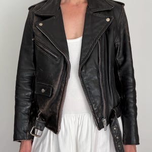 May include: Black leather motorcycle jacket with a classic asymmetrical zip closure, featuring silver hardware, a notched collar, and multiple zippered pockets. The jacket has a worn, distressed appearance, with a belt at the waist.