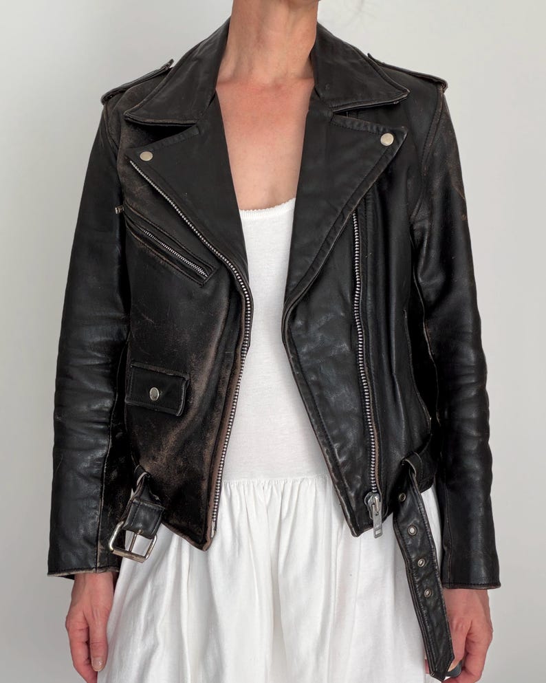 May include: Black leather motorcycle jacket with a classic asymmetrical zip closure, featuring silver hardware, a notched collar, and multiple zippered pockets. The jacket has a worn, distressed appearance, with a belt at the waist.
