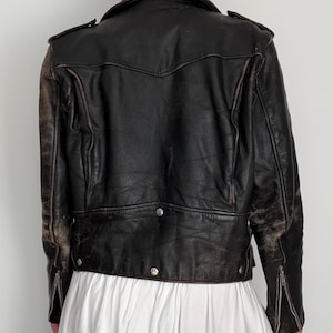 May include: A vintage black leather jacket with a distressed appearance. The jacket features a notched collar, shoulder epaulets, and silver-toned hardware. Zippers adorn the sleeves. The back of the jacket has a decorative yoke and a buttoned hem.