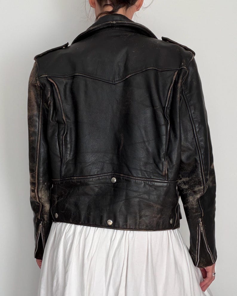 May include: A vintage black leather jacket with a distressed appearance. The jacket features a notched collar, shoulder epaulets, and silver-toned hardware. Zippers adorn the sleeves. The back of the jacket has a decorative yoke and a buttoned hem.