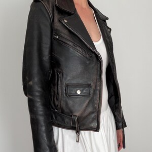 May include: A distressed black leather motorcycle jacket with a notched collar, asymmetrical zipper, and multiple pockets. The jacket features a belt at the waist and silver hardware. The leather shows signs of wear, giving it a vintage look.