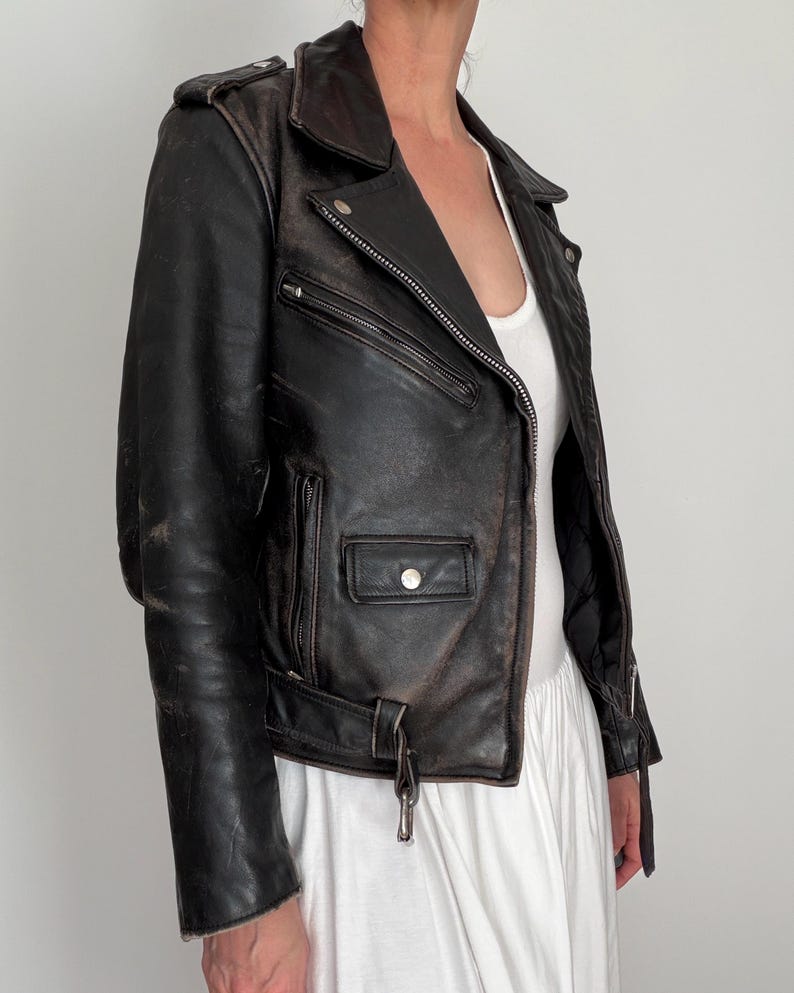 May include: A distressed black leather motorcycle jacket with a notched collar, asymmetrical zipper, and multiple pockets. The jacket features a belt at the waist and silver hardware. The leather shows signs of wear, giving it a vintage look.