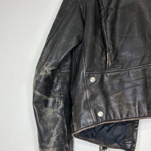 May include: Close-up of a distressed black leather jacket. The jacket features a classic design with silver snap closures and a zipper. The leather shows signs of wear, adding to its vintage appeal. The lining is black.