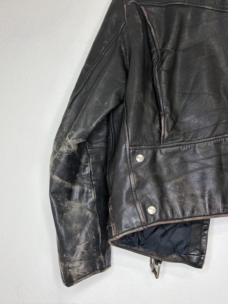 May include: Close-up of a distressed black leather jacket. The jacket features a classic design with silver snap closures and a zipper. The leather shows signs of wear, adding to its vintage appeal. The lining is black.