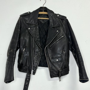 May include: Black leather motorcycle jacket with a notched collar, asymmetrical zipper closure, and multiple pockets. The jacket features a belt at the waist and is hanging on a wooden hanger. The interior lining is quilted.