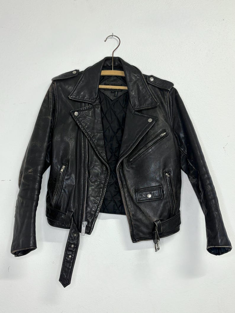 May include: Black leather motorcycle jacket with a notched collar, asymmetrical zipper closure, and multiple pockets. The jacket features a belt at the waist and is hanging on a wooden hanger. The interior lining is quilted.