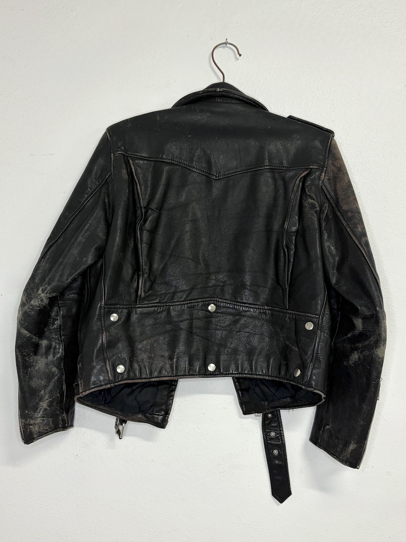 May include: Black leather jacket with silver hardware, hanging on a metal hanger. The jacket has a classic biker style with a notched collar, asymmetrical zipper, and multiple pockets. The leather shows signs of wear.