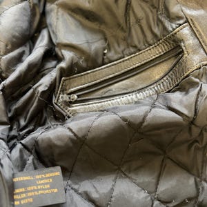 May include: Close-up of the interior of a black leather jacket. The quilted lining is visible, along with a zippered pocket. A tag indicates the outer shell is 100% genuine leather, the lining is 100% nylon, and the filler is 100% polyester.