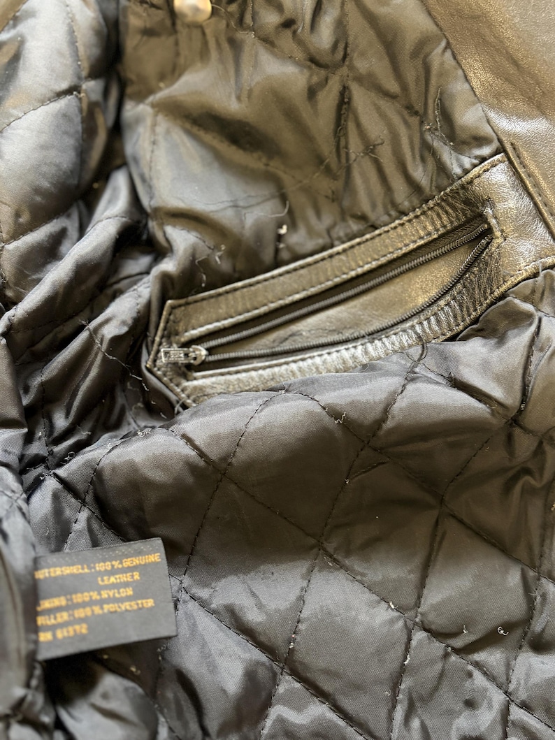 May include: Close-up of the interior of a black leather jacket. The quilted lining is visible, along with a zippered pocket. A tag indicates the outer shell is 100% genuine leather, the lining is 100% nylon, and the filler is 100% polyester.
