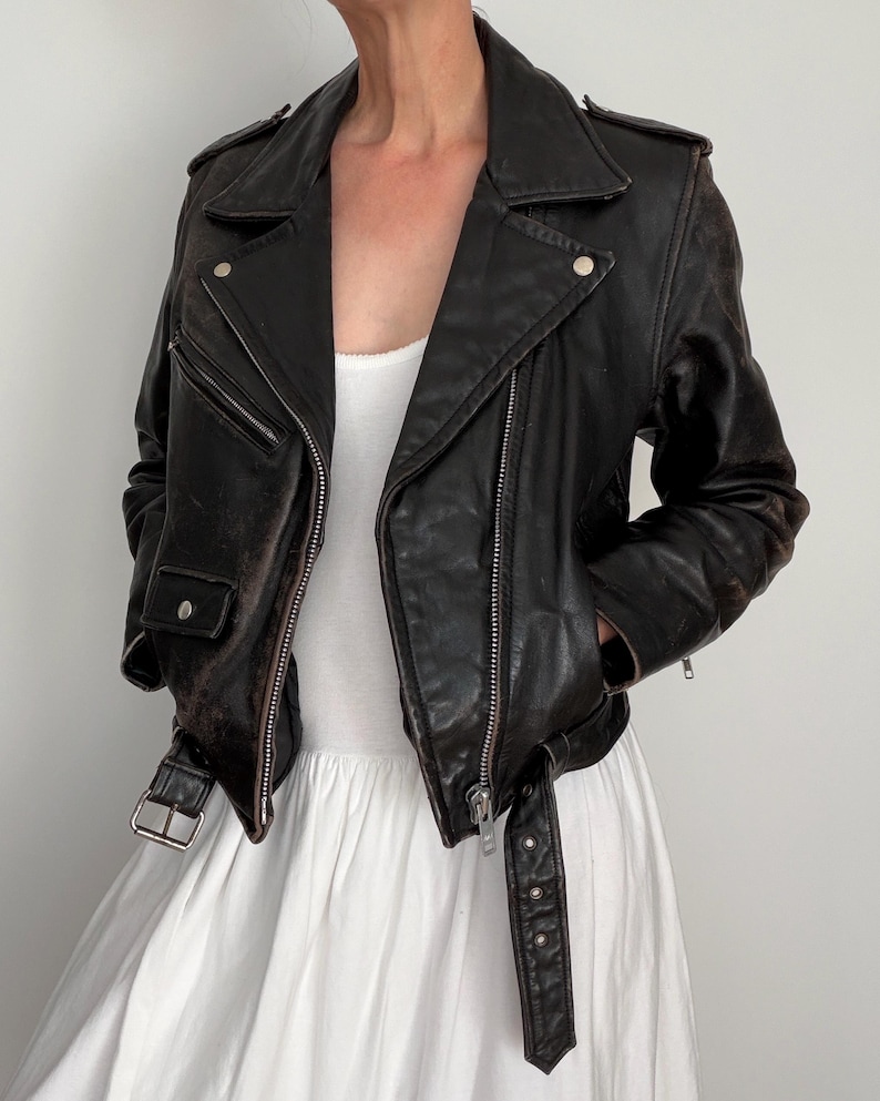 May include: A black leather motorcycle jacket with a classic asymmetrical zipper design. The jacket features a notched collar, silver hardware, and a belt at the waist. The leather appears worn, giving it a vintage aesthetic.