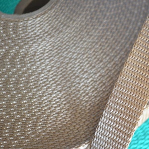 May include: Close-up of a roll of beige woven ribbon. The ribbon has a textured, diamond-patterned weave. A length of the ribbon extends from the roll, showing the same pattern. The background is a mix of beige and teal.