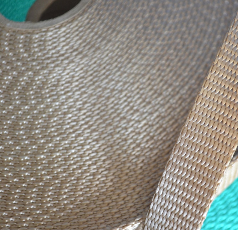 May include: Close-up of a roll of beige woven ribbon. The ribbon has a textured, diamond-patterned weave. A length of the ribbon extends from the roll, showing the same pattern. The background is a mix of beige and teal.