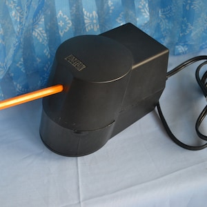 May include: Black electric pencil sharpener with a cord and an orange pencil sticking out of the sharpening hole. The sharpener has the word "BRAXTON" printed on the side.