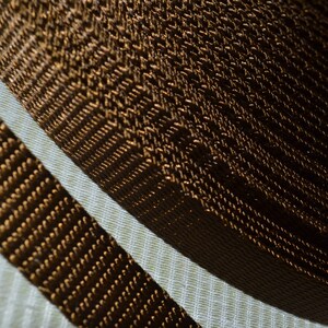May include: Close-up of a roll of brown woven webbing, with a parallel strip of the same material. The webbing has a textured, ribbed appearance, and the color is a warm, dark brown. The background is a light, textured fabric.