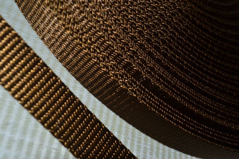 May include: Close-up of a roll of brown woven webbing, with a parallel strip of the same material. The webbing has a textured, ribbed appearance, and the color is a warm, dark brown. The background is a light, textured fabric.