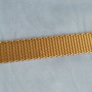 May include: Close-up of a golden-yellow woven strap. The strap has a textured, ribbed surface and is set against a light blue fabric background. The strap appears to be made of a durable material.