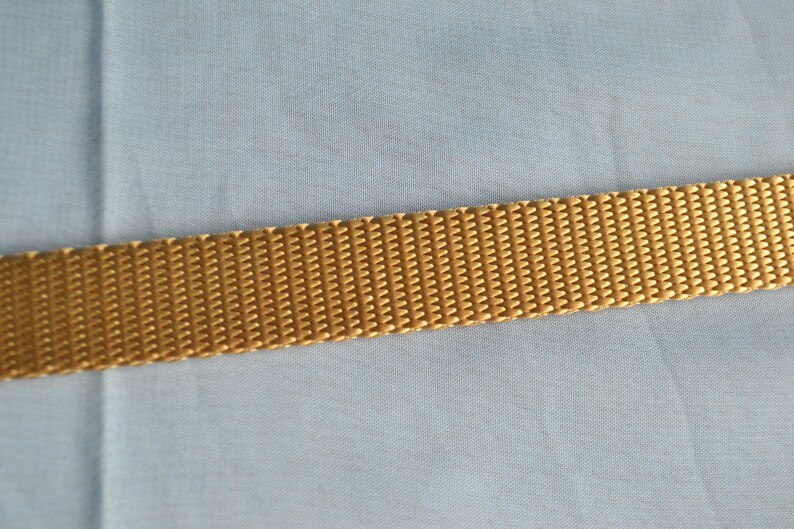 May include: Close-up of a golden-yellow woven strap. The strap has a textured, ribbed surface and is set against a light blue fabric background. The strap appears to be made of a durable material.