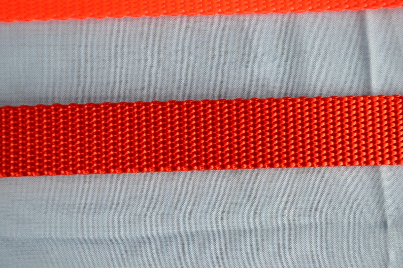 May include: Close-up of two bright orange nylon webbing straps. The top strap is thinner than the bottom strap. The straps are set against a light blue fabric background. The straps are likely used for crafting or outdoor gear.
