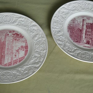 May include: Two white ceramic plates with raised designs. The plates feature pink images of buildings. One plate shows a church with a tall spire, the other shows a building with multiple windows.