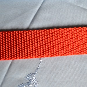 May include: Close-up of a bright orange woven strap. The strap is made of a tightly woven material with a textured surface.