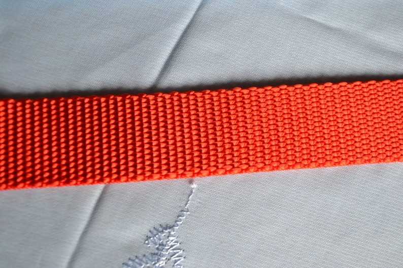May include: Close-up of a bright orange woven strap. The strap is made of a tightly woven material with a textured surface.