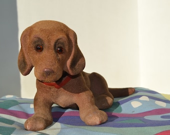 May include: A brown plush toy dog with red eyes and a red collar. The dog is lying on a colorful patterned fabric.