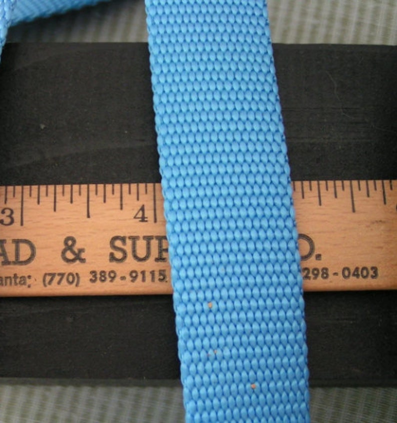May include: A close-up of a light blue webbing strap, measuring 1 inch wide, against a black surface and a wooden ruler. The ruler has the text "AD & SUP" and a phone number.