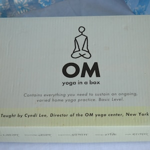 May include: A light blue box with the text "OM yoga in a box" and a seated figure logo. The box contains everything needed for a home yoga practice. The text on the box says it is taught by Cyndi Lee, Director of the OM yoga center, New York City.