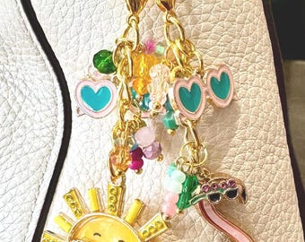 Könnte beinhalten: Ein goldfarbener Schlüsselanhänger-Anhänger mit einer lächelnden Sonne mit Sonnenbrille, einem rosa Flamingo mit Sonnenbrille und herzförmigen Sonnenbrillen. Der Anhänger ist mit bunten Perlen verziert und hängt an einer goldenen Kette.